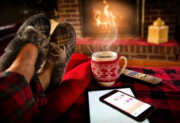 Füße in Hausschuhen vor dem Kaminfeuer, daneben eine heiße Tasse Kaffee und ein Smartphone